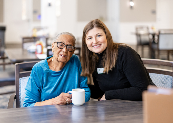 Smiling senior resident in a blue sweater enjoys a warm beverage while sitting with a friendly staff member who is wearing a black sweater, showcasing the warmth and companionship in the community.