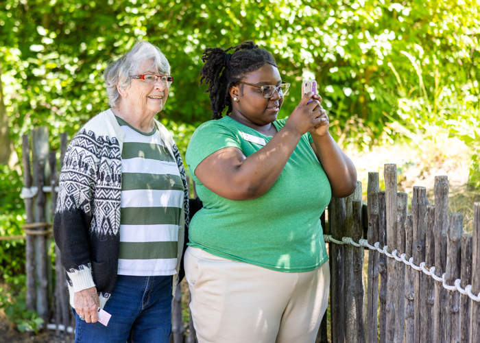 Two women enjoying a sunny day outdoors, with one taking a photo on her phone and the other smiling beside her, standing by a wooden fence surrounded by lush greenery.