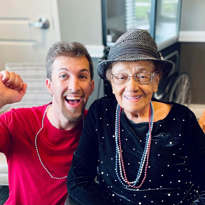Senior resident in a festive hat and beads smiles alongside a cheerful young man in a red shirt, both celebrating together at a Fourth of July event in a senior living community.