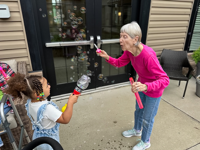 A senior resident in a pink shirt and a young girl with curly hair joyfully blow bubbles together outdoors, sharing a playful and heartwarming moment across generations.
