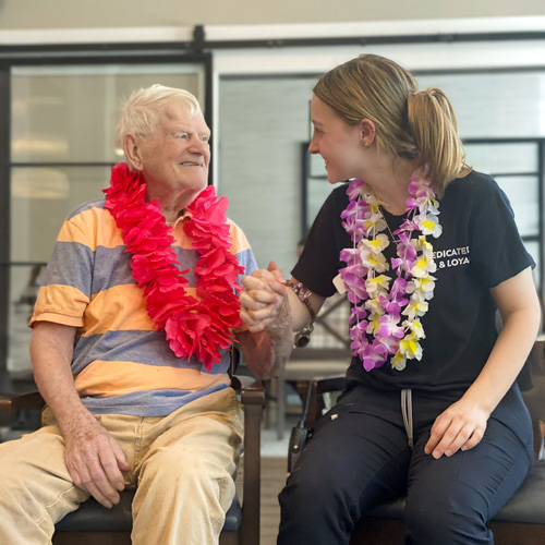 Senior resident wearing a red lei holds hands with a staff member in a purple lei, sharing a happy moment together, emphasizing the supportive community environment.