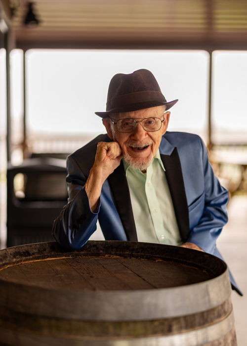 Senior resident in a sharp blue blazer and hat, smiling joyfully while resting his arm on a wooden barrel, celebrating a special outdoor event at a senior living community.
