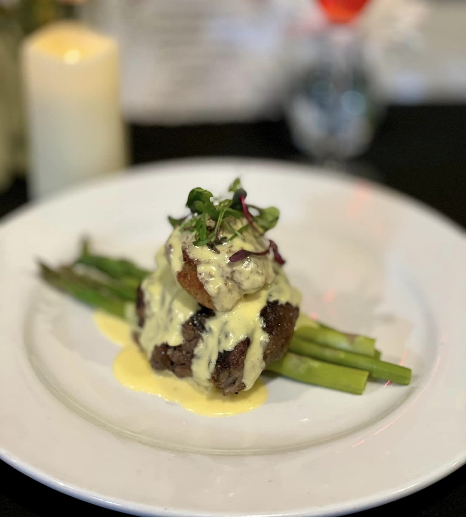 Plated gourmet steak with asparagus, topped with creamy hollandaise sauce and garnished with microgreens, set against a blurred background of a candle, flowers, and drink.