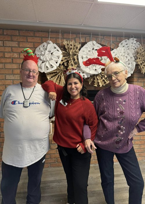 Senior Living residents and employee pose together while wearing Christmas headbands while paper snowflakes decorate the background.