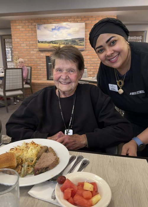 Walden Place Senior Living resident and employee pose together in the dining area with a delicious balanced meal on the table.