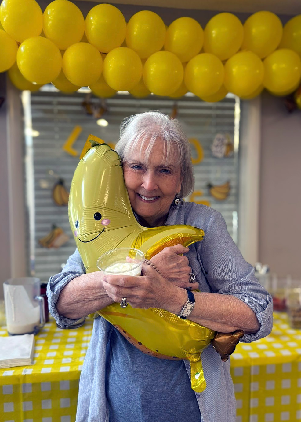 A senior woman smiles, clutching a balloon shaped like a happy banana, with banana themed decor in the background.