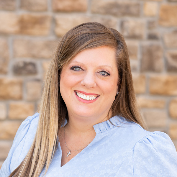 Katie Max, Senior Living Director at The Henry Senior Living, smiling in a professional headshot with long highlighted light brown hair, a light blue blouse, a cross necklace, and a tan stone wall background.