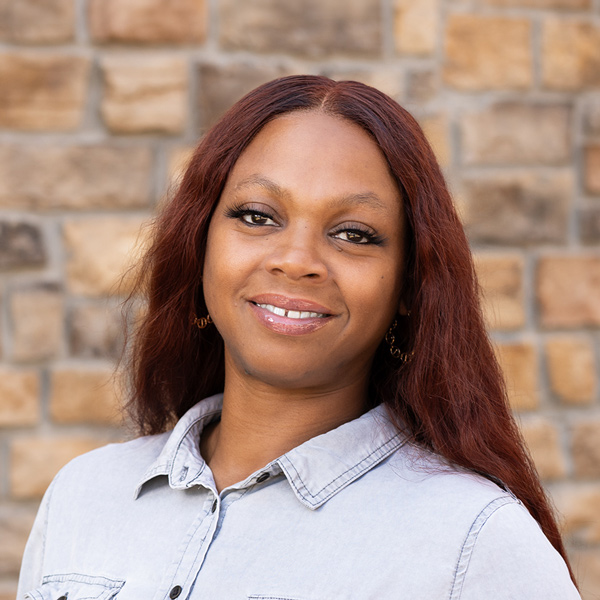 Kimbra Turner, Memory Care Director at The Henry Senior Living, looking at the camera in a professional headshot with long auburn hair, gold hoop earrings, a light gray button-up shirt, and a tan stone wall background.