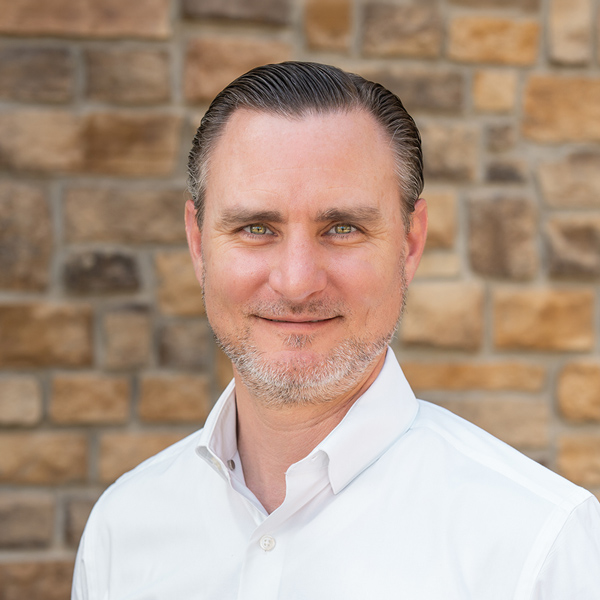 Kris Schroeder, Executive Director at The Henry Senior Living, looking at the camera in a professional headshot with slicked-back dark hair, a short salt-and-pepper beard, a white button-up shirt, and a tan stone wall background.
