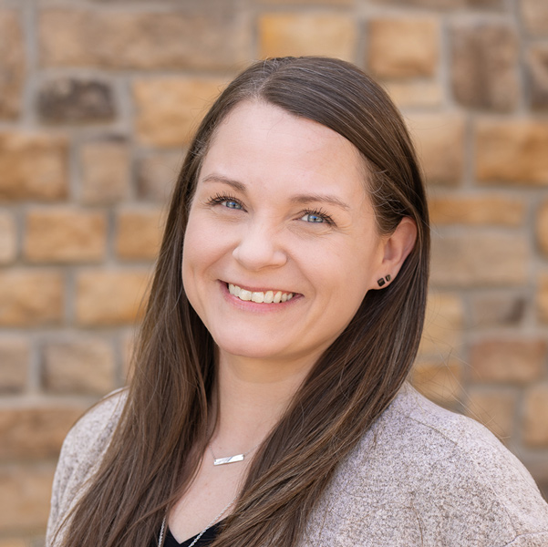 Lauren Stenger, Resident Services Director at The Henry Senior Living, smiling in a professional headshot with long straight brown hair, small black stud earrings, a layered necklace, and a tan stone wall background.