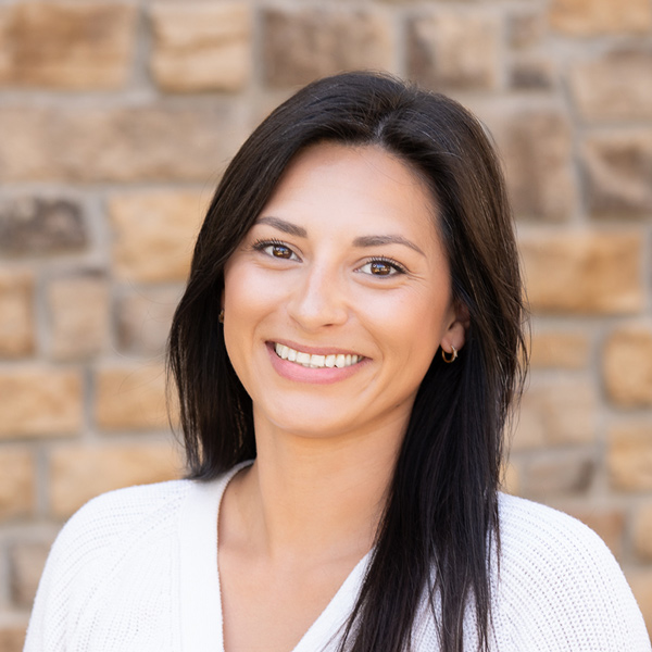 Liz Buneta, Business Office Director at The Henry Senior Living, smiling in a professional headshot with long dark hair, small hoop earrings, a white cardigan, and a tan stone wall background.