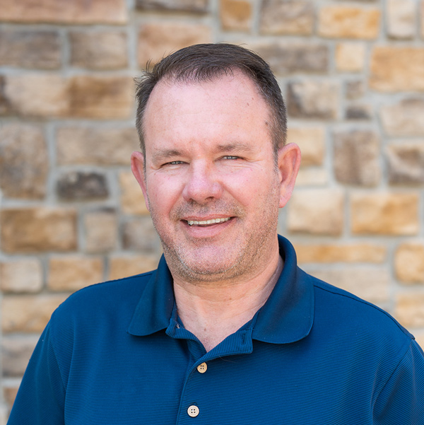 Scott Hall, Plant Operations Director at The Henry Senior Living, looking at the camera in a professional headshot with short brown hair, a blue polo shirt, and a tan stone wall background.