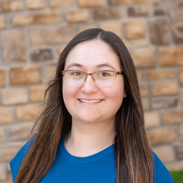 Emilee Luck, Wellness Director at The Henry Senior Living, smiling in a professional headshot with long dark hair, glasses, a bright blue top, and a tan stone wall background.