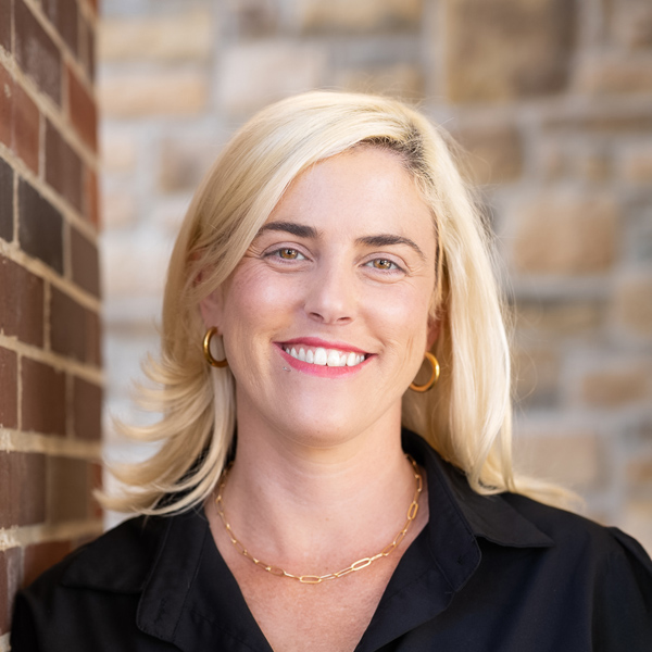 Michelle Graham, Arrow Wellness Director at Arrow Senior Living Home Office providing Executive Director interim support for The Henry Senior Living, smiling in a professional headshot with blonde hair, gold hoop earrings, a black blouse, and a softly blurred brick and stone background.