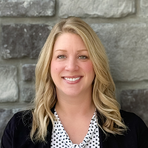 Amanda Bernhardt, Senior Living Director at The Henry Senior Living, smiling in front of a gray stone wall, wearing a polka-dot blouse and black cardigan.